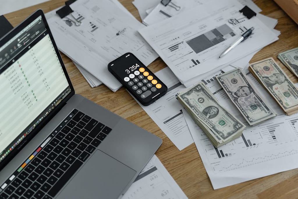 'Desk with financial documents, a laptop showing a spreadsheet, a smartphone with a calculator app displaying 3254, and stacks of U.S. dollar bills.'