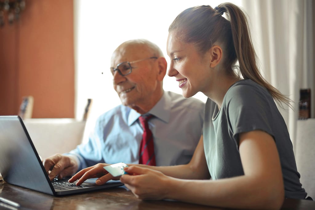A woman and an older man sitting at a table with a laptop, engaged in a conversation and working together.