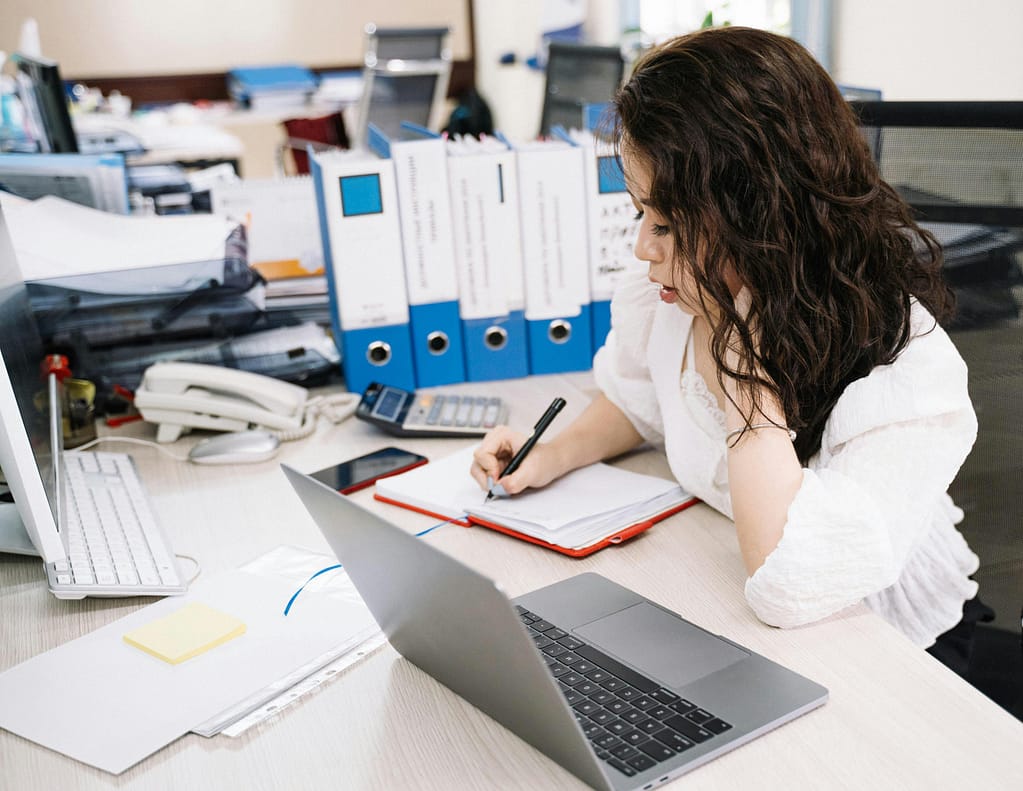 Woman in white shirt, seated at a desk, concentrating while writing in a notebook. A laptop, phone, and office supplies are on the desk, with file binders and a printer in the background.